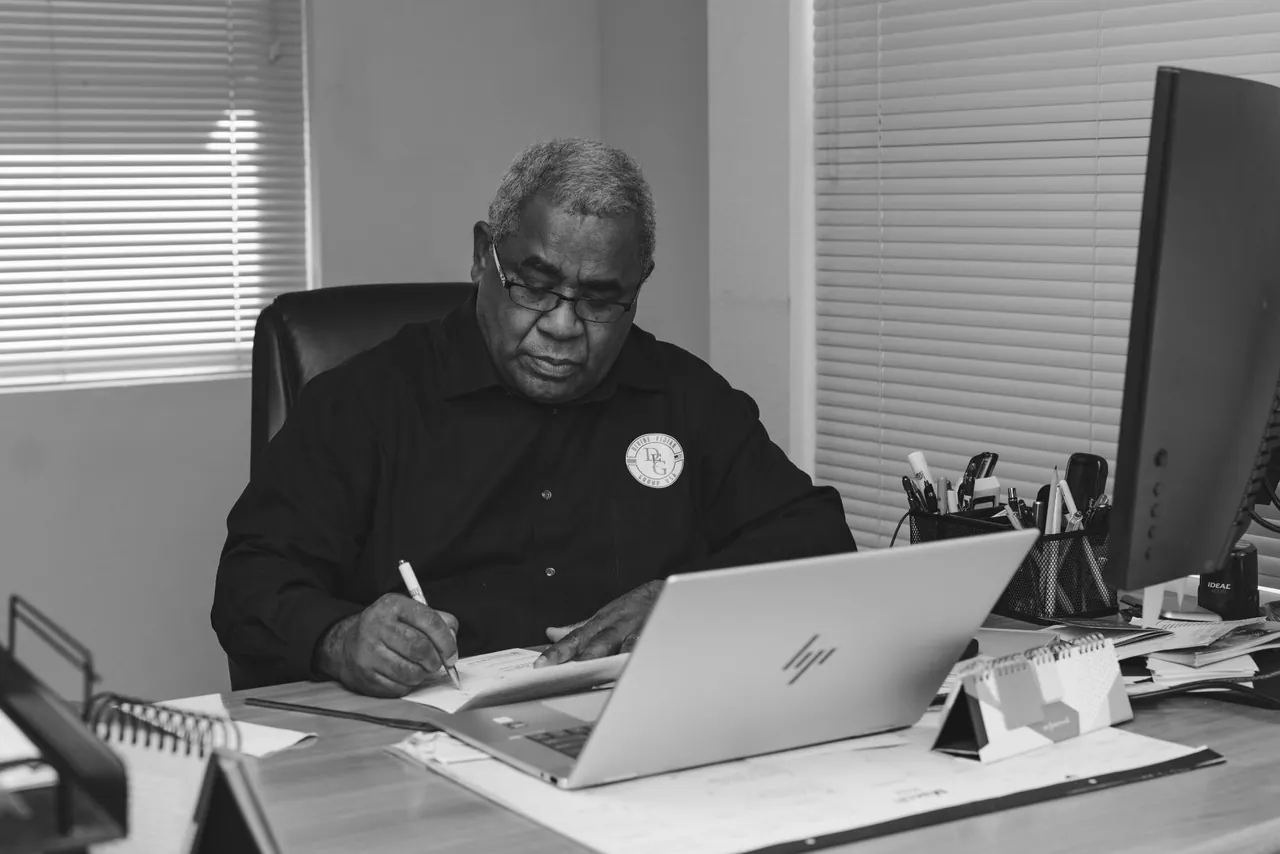 A man writing at an office desk beside a laptop.